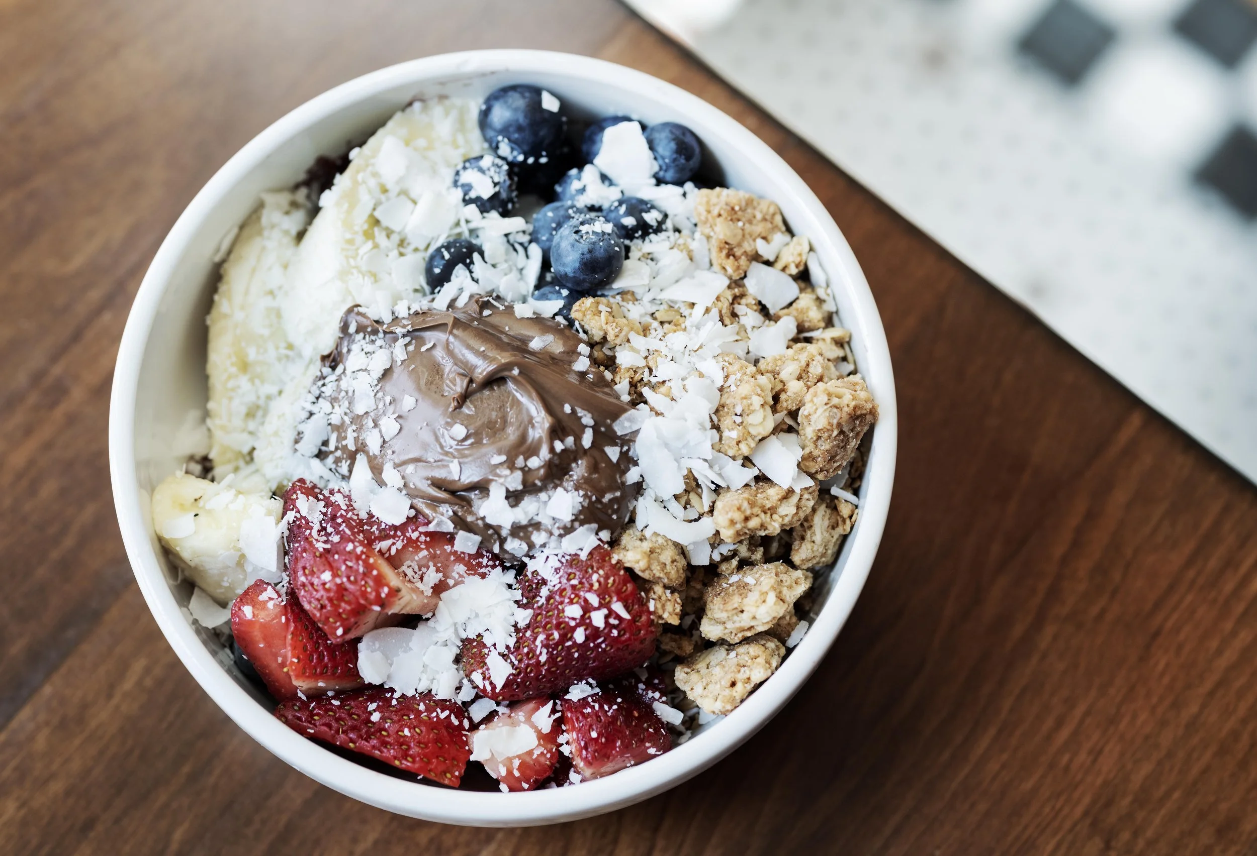 A close-up of a bowl of mixed frozen yogurt topped with blueberries, sliced strawberries, granola, white chocolate shavings, and a dollop of chocolate spread, sitting on a wooden surface.
