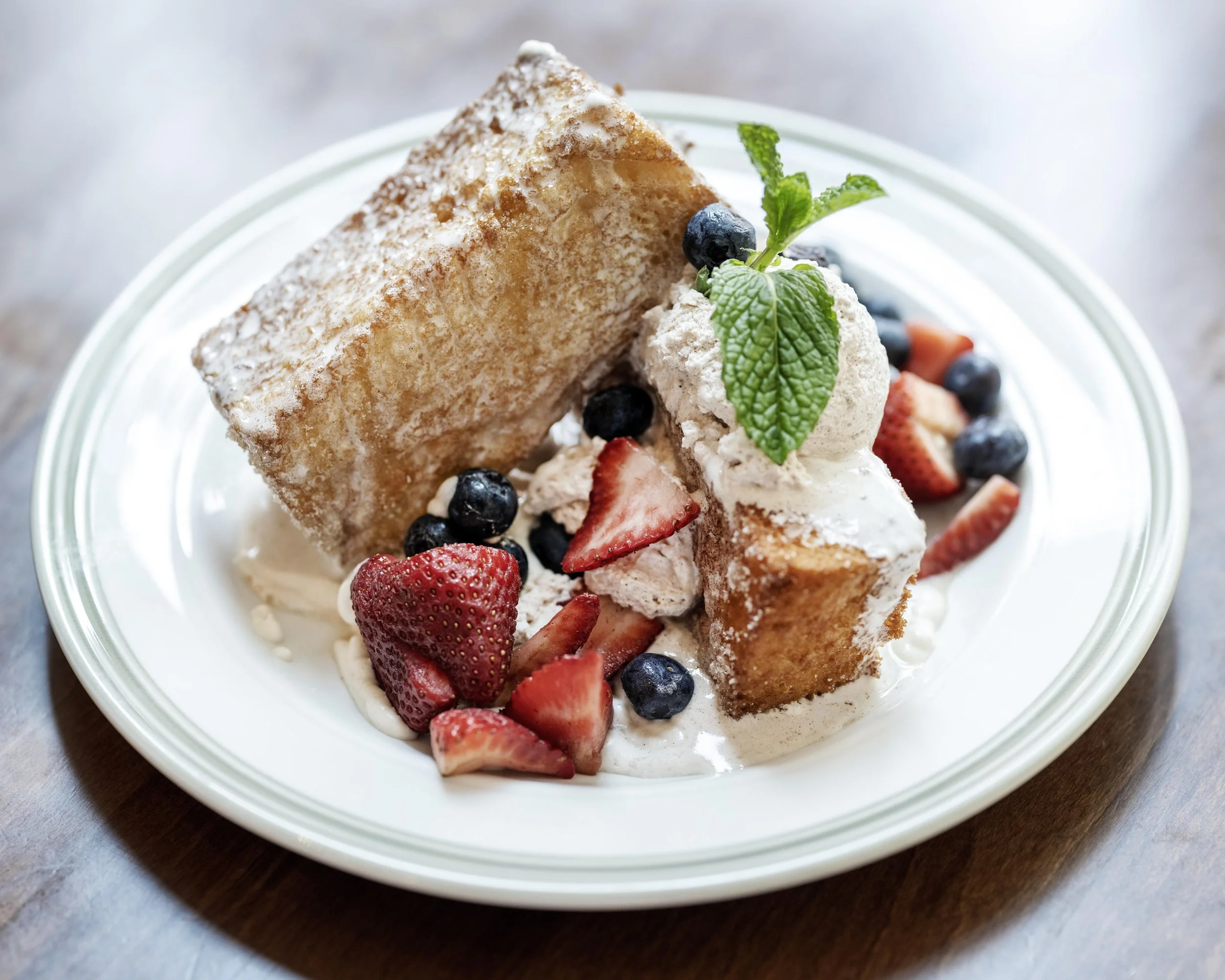 Dessert with fried bread, whipped cream, strawberries, blueberries, and a mint leaf on a white plate.