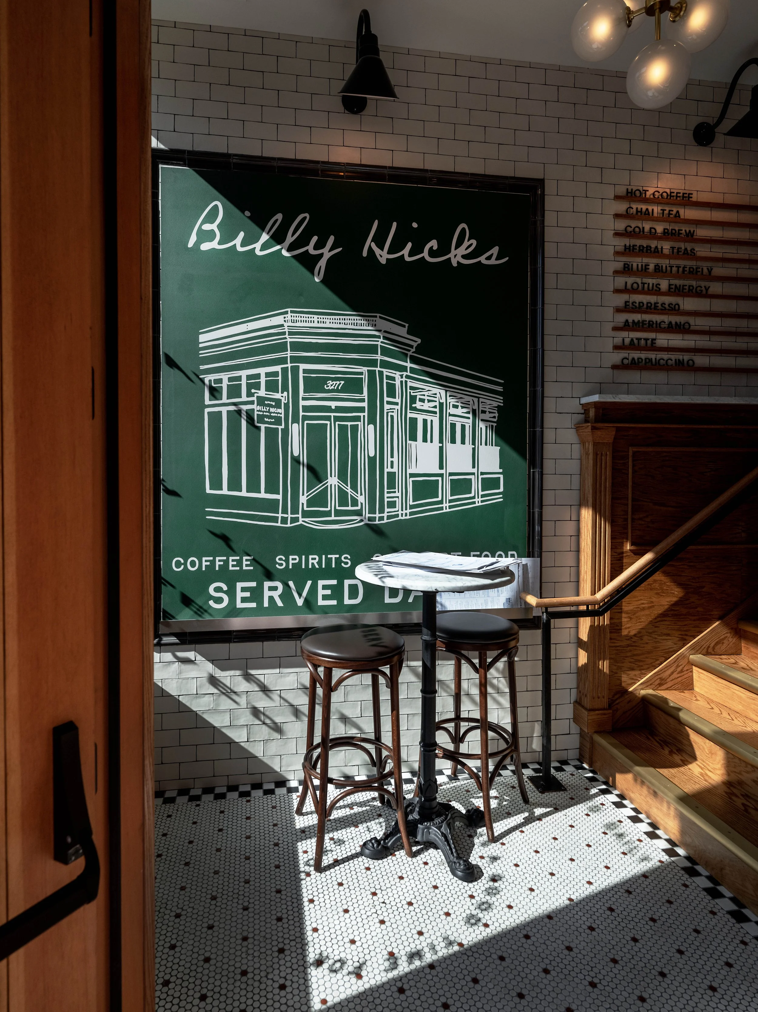Interior of a cafe with a chalkboard sign reading "Billy Hicks" featuring a line drawing of a building, a small round table with two bar stools, and a menu board on the wall listing coffee offerings.