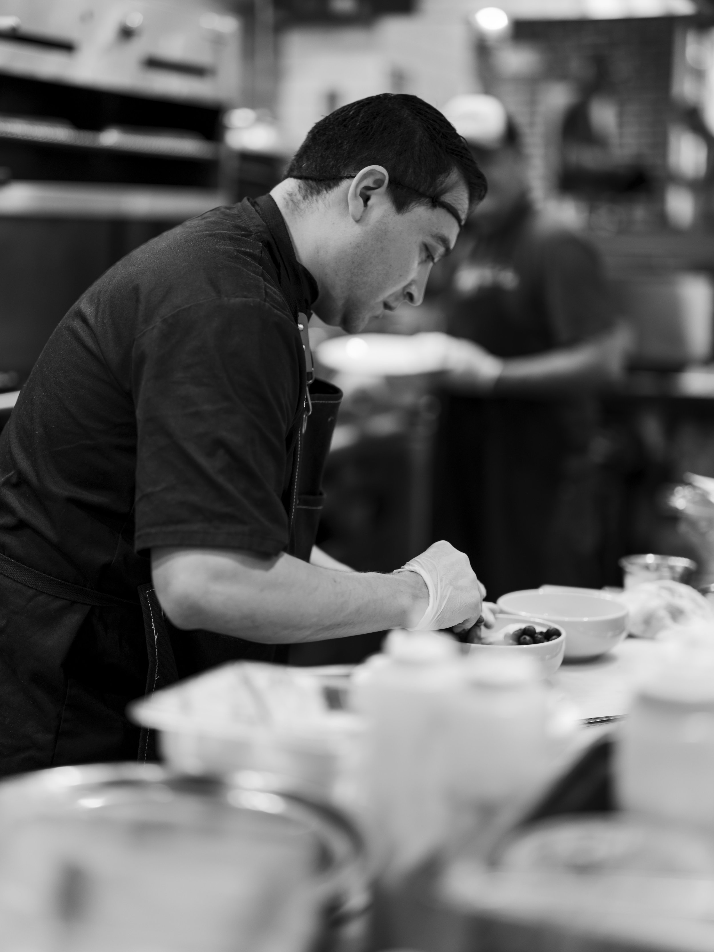 A chef preparing food in a professional kitchen, wearing gloves, with bowls and kitchen utensils around him.