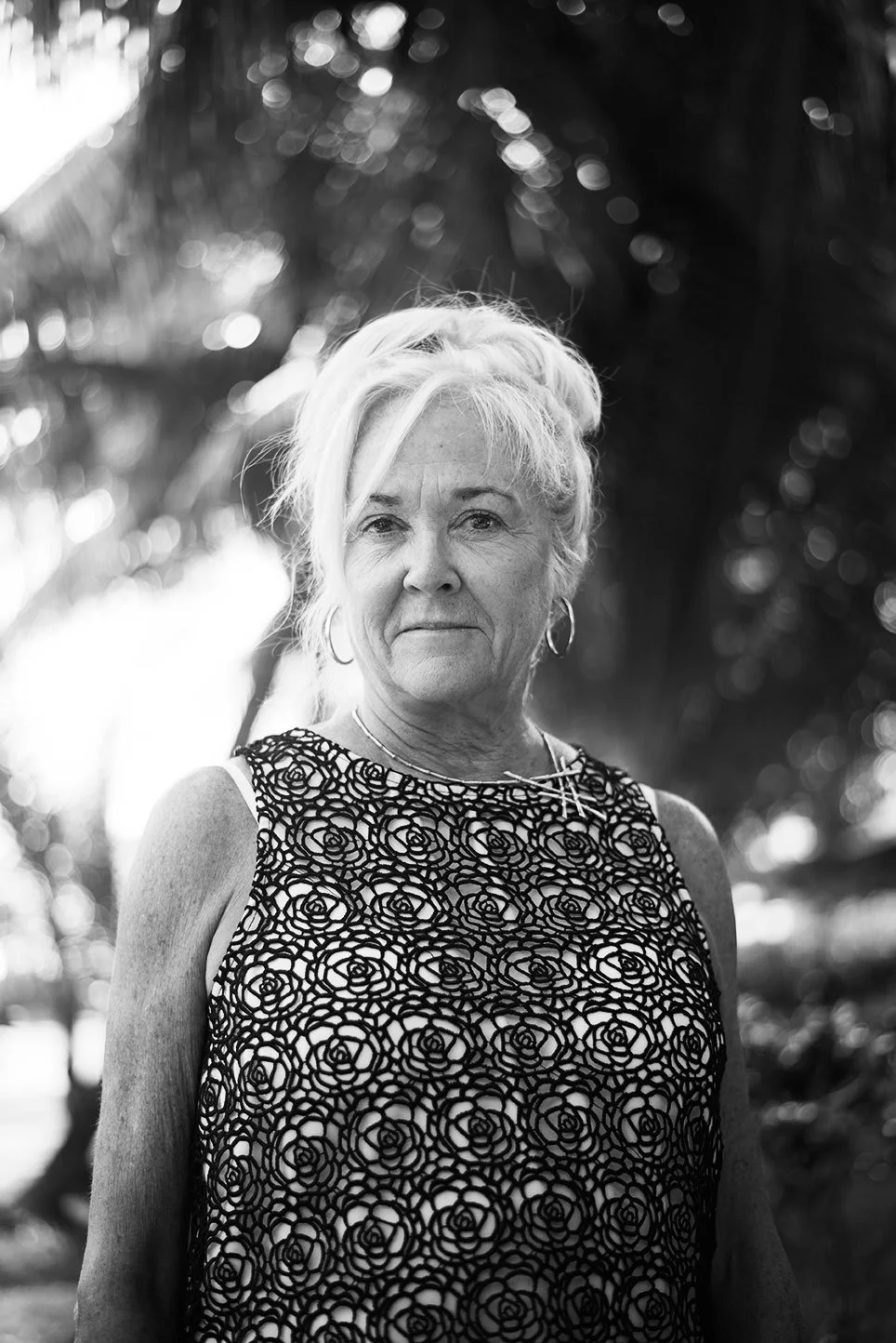 Black and white photo of an elderly woman with short, light-colored hair, wearing a sleeveless top with a floral pattern, standing outdoors with blurred trees in the background.