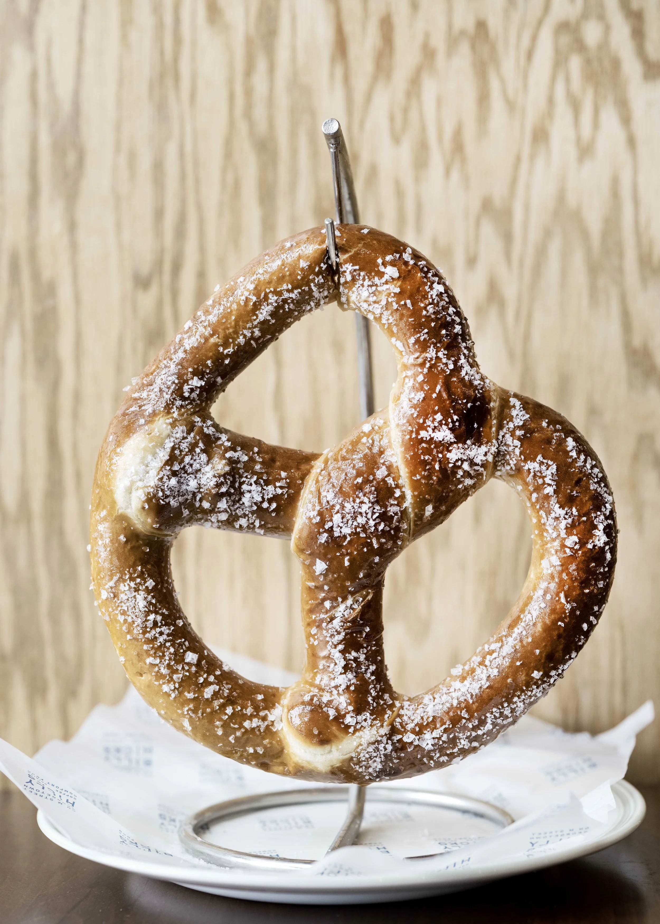 A large salted pretzel on a metal stand, served on a white plate with a paper liner, against a wooden background.