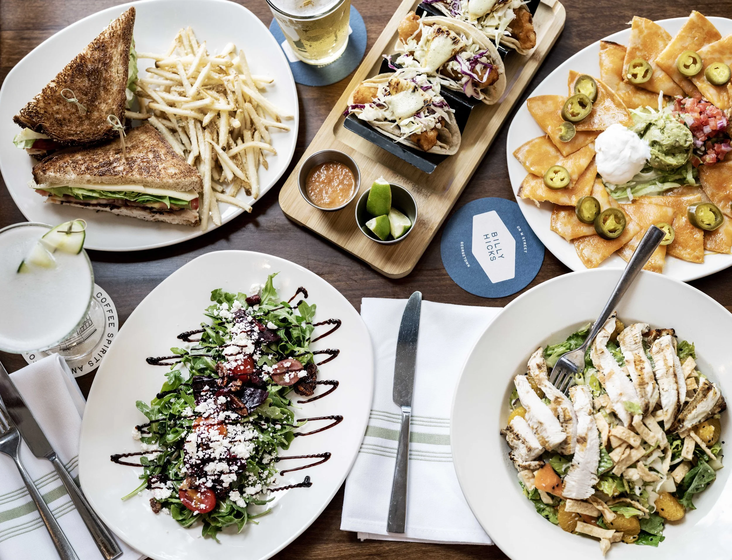 A variety of dishes on a wooden table including a Club sandwich with french fries, a taco platter with lime wedges, a nacho platter with jalapeños and guacamole, a grilled chicken salad, and a mixed greens salad with cherry tomatoes and grated cheese