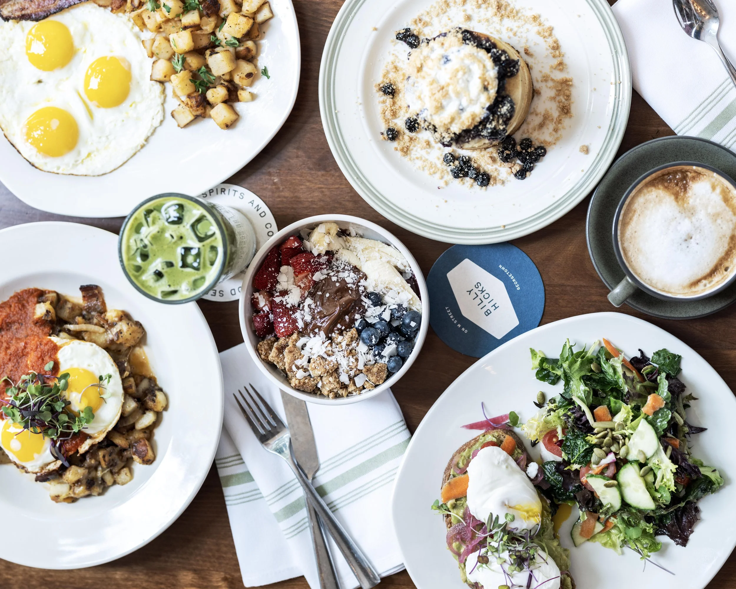 A spread of breakfast foods including sunny side up eggs, potatoes, a berry parfait, a green drink, a salad with poached eggs, a cappuccino, and a dessert with blueberries.