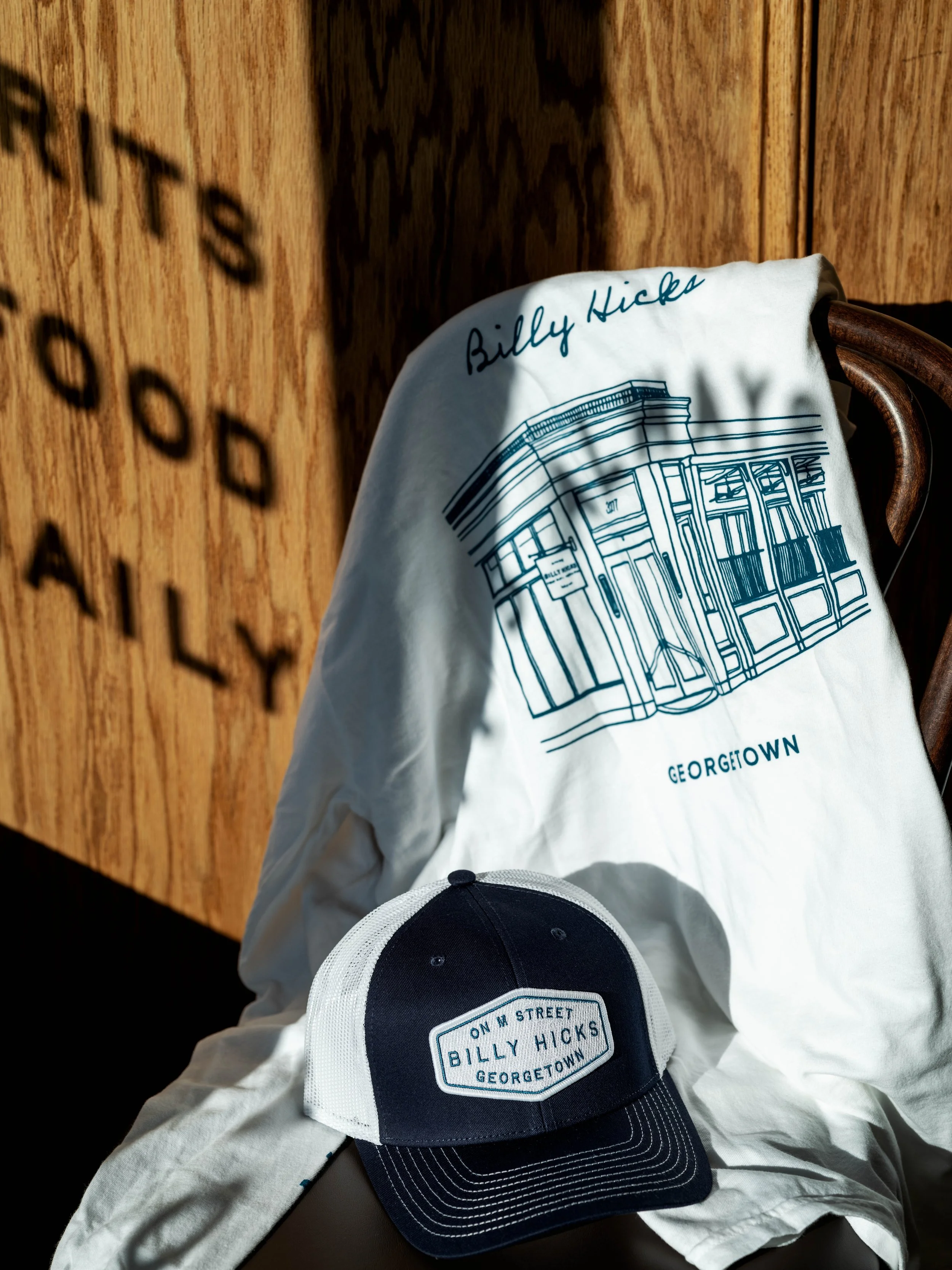 A white T-shirt with blue print of a building and text reading 'Billy Hicks' and 'Georgetown', hangs on a wooden chair. In front of it, a navy and white cap with a patch reading 'On M Street Billy Hicks Georgetown' rests on a white cloth.