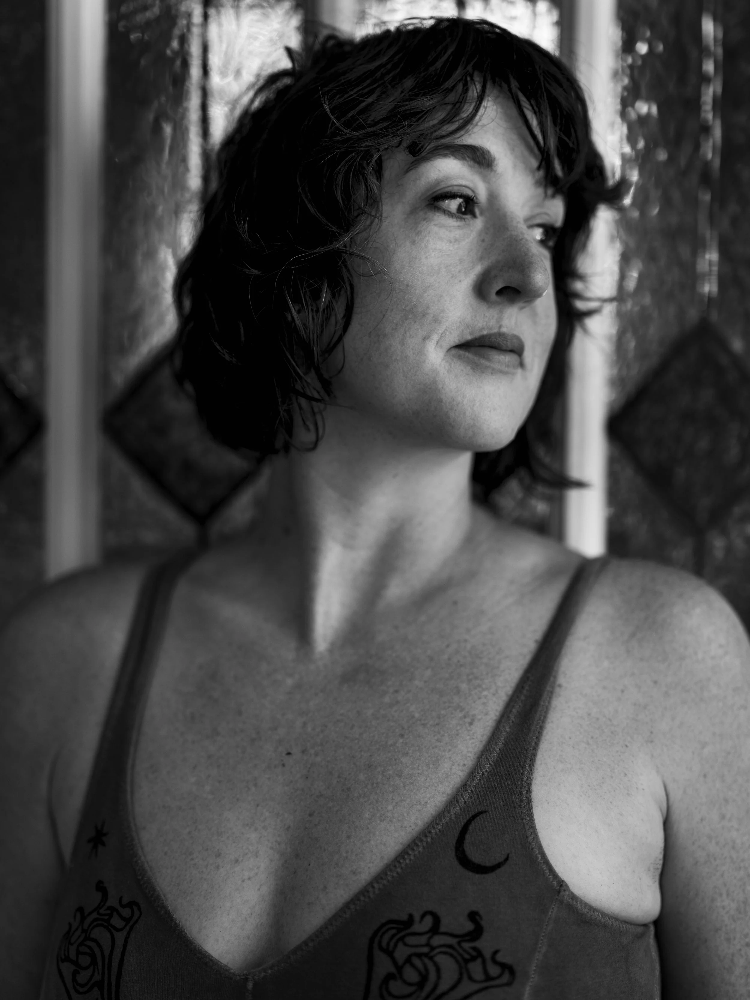 A woman with short curly hair looking thoughtfully to the side, wearing a tank top with a moon and dragon tattoo, standing in front of a wooden fence.