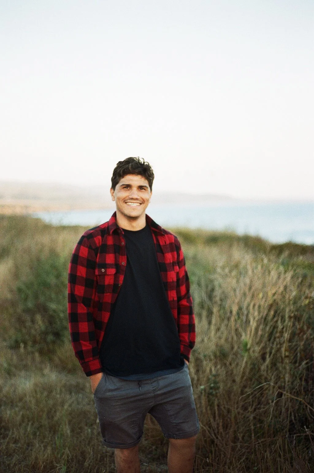 A young man with dark hair standing outdoors by the coast, smiling, wearing a black t-shirt, a red and black plaid shirt, and gray shorts, with grassy dunes and the ocean in the background on a clear day.