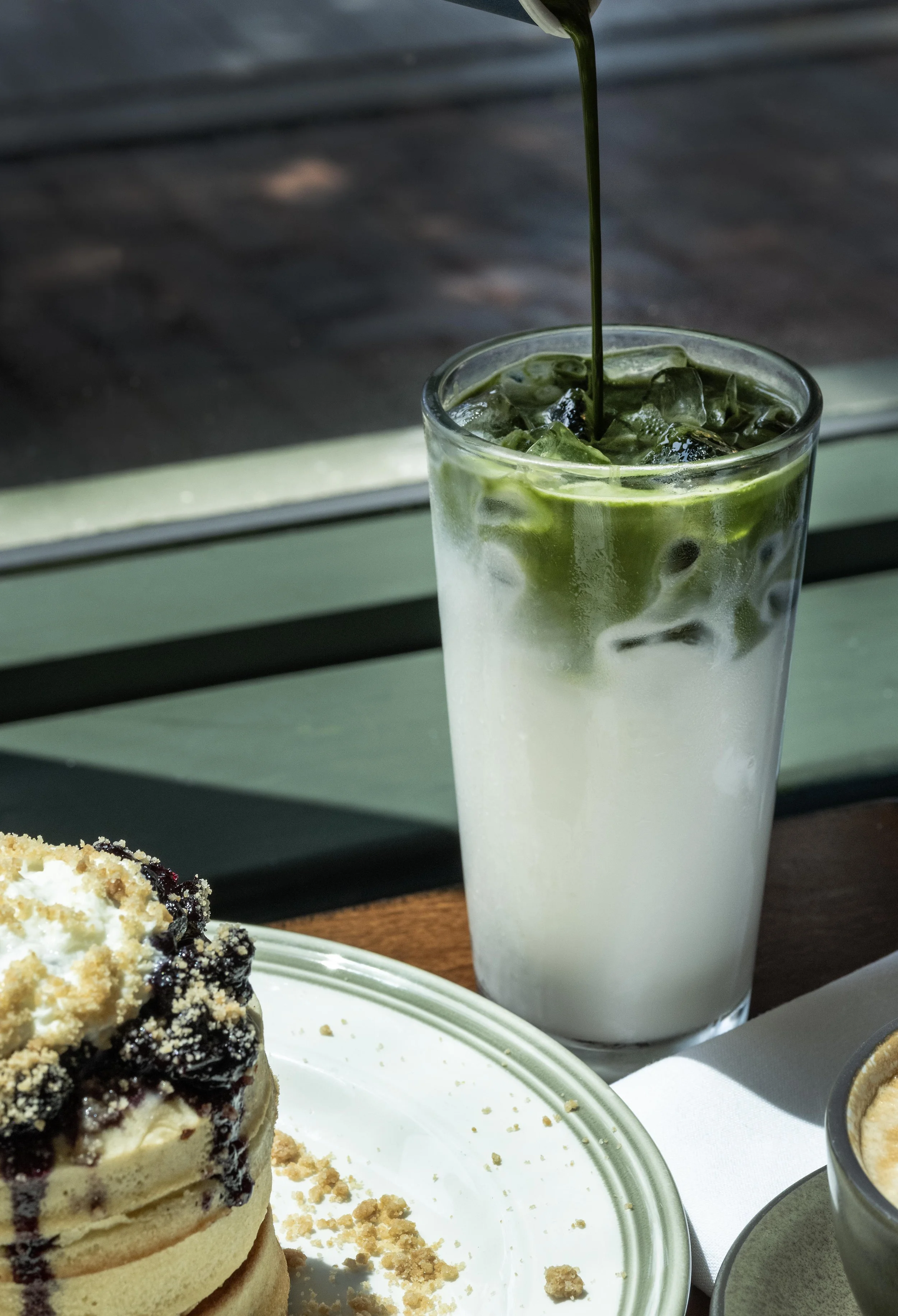 Glass of iced matcha latte with milk, served with a green straw, on a table next to a dessert.
