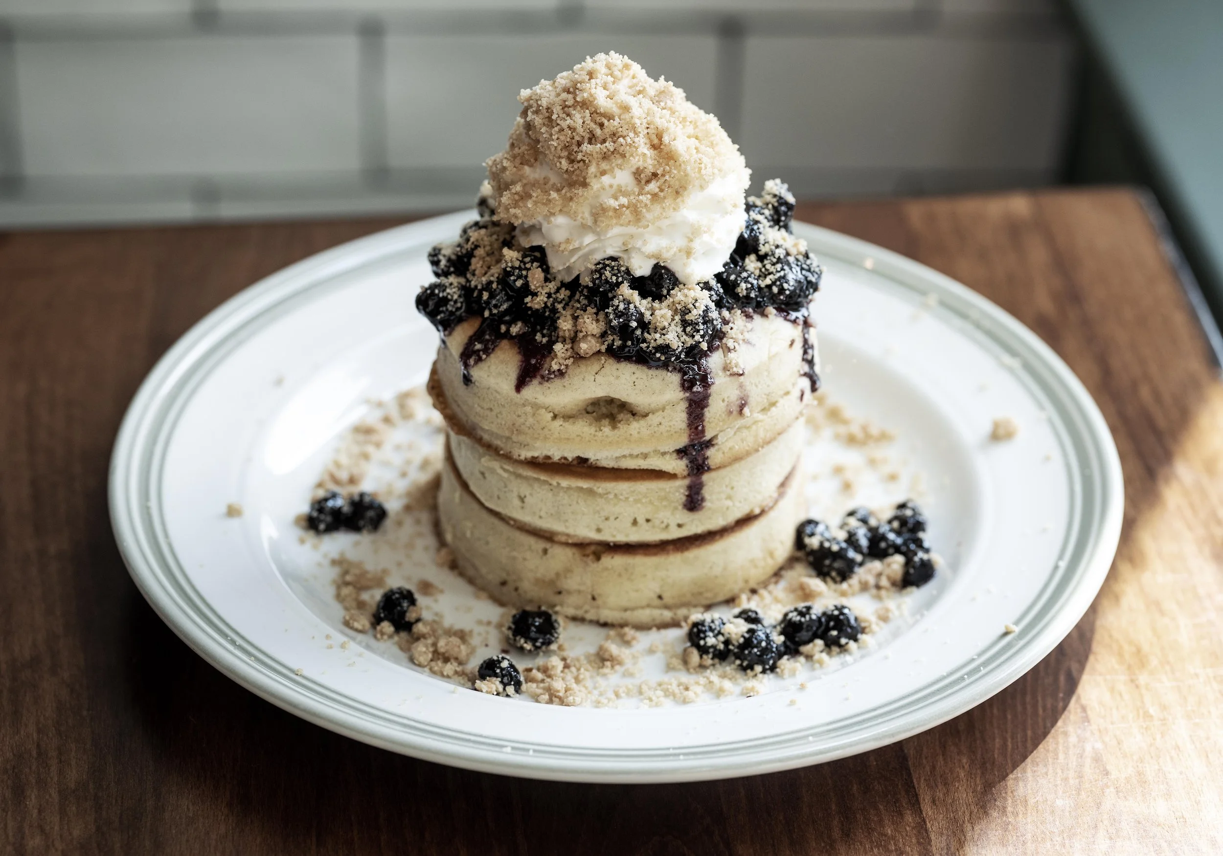 Stack of three blueberry matcha pancakes topped with whipped cream, blueberry sauce, powdered crumbs, and a scoop of ice cream on a white plate with scattered crumbs and blueberries.