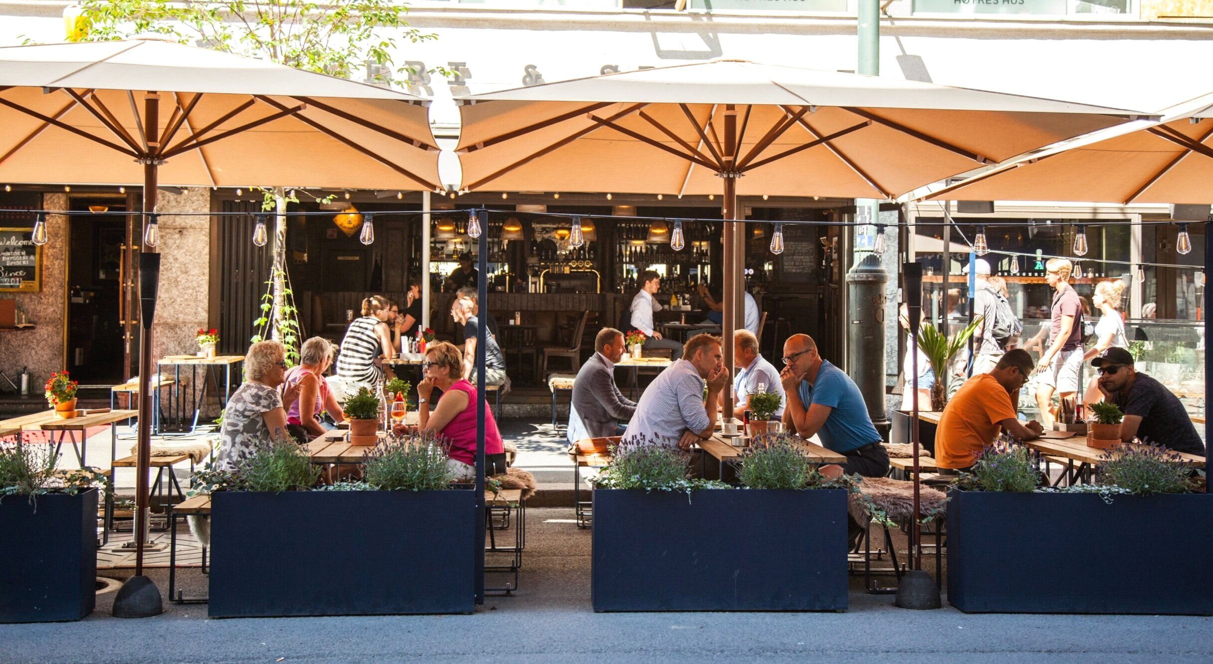 Uteservering med folk som spiser og prater under store beige parasoller utenfor en bar eller restaurant