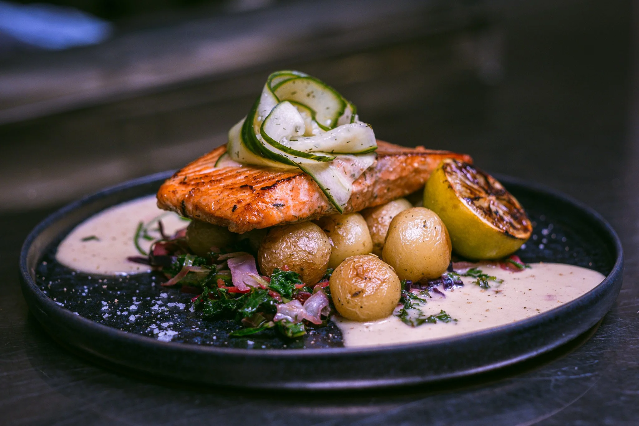 Grillet laks med sitron, poteter, og grønnsaker på en svart tallerken.