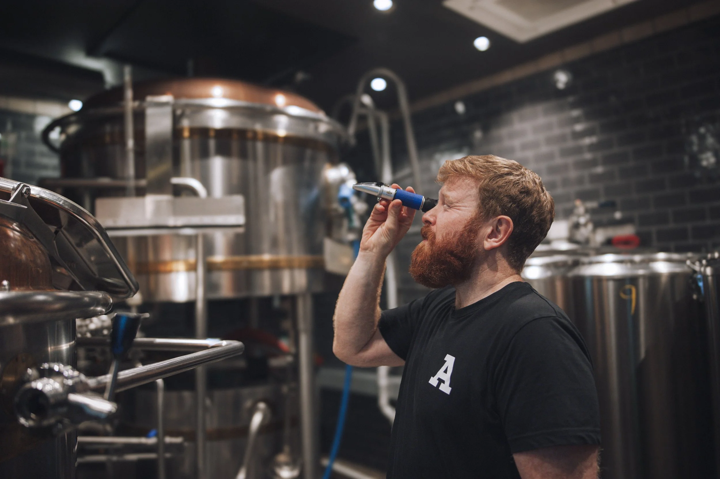 En mann med rød beard og svart t-skjorte ser gjennom et mikroskop i et industrielt kjøkken eller bryggeri med store metallkummer.