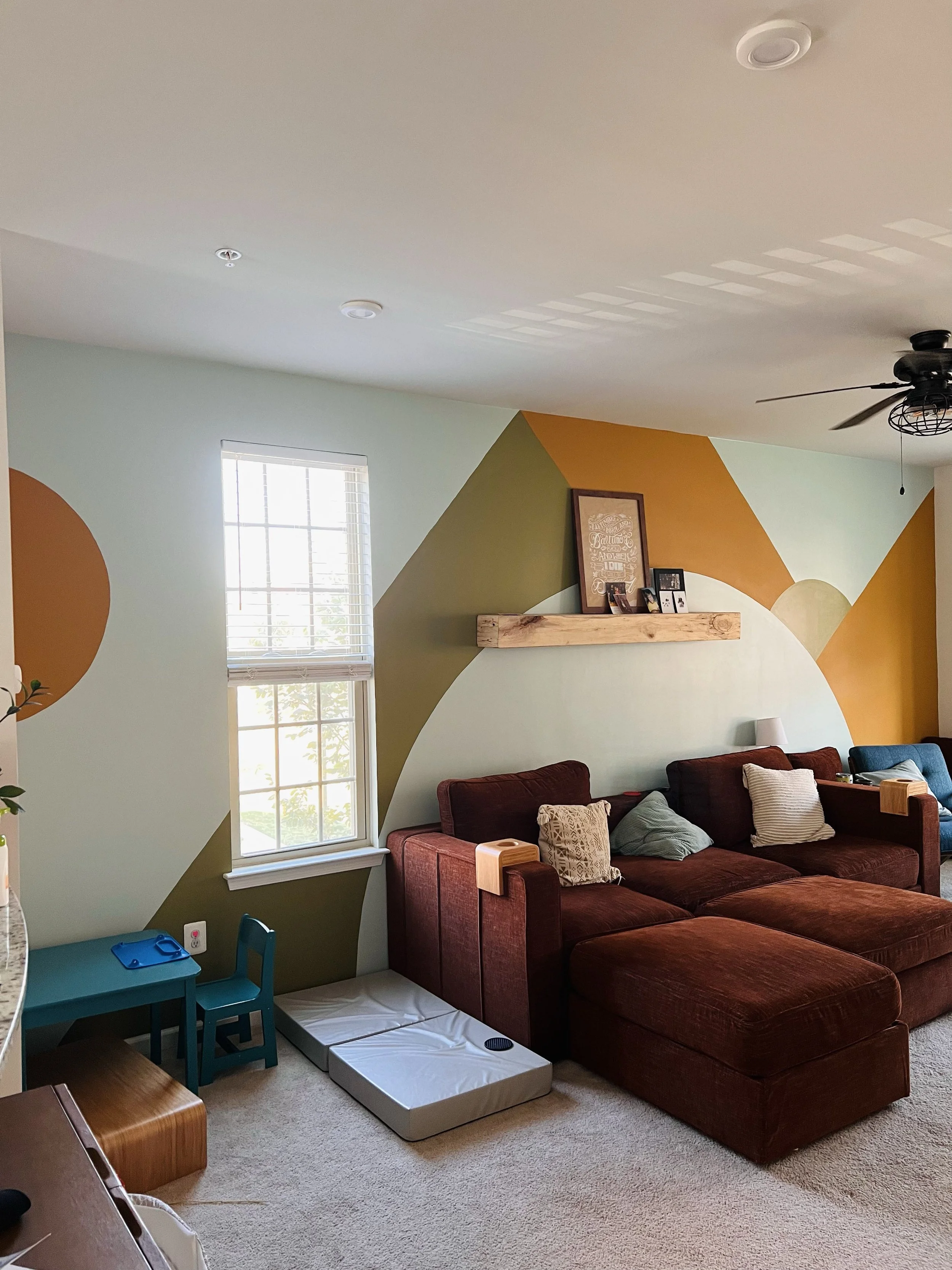 A living room with light green walls featuring geometric accents in orange, dark green, and cream. There is a large red sectional sofa with decorative pillows, a small blue table and chairs, a window with blinds, a ceiling fan, and a wooden shelf wit