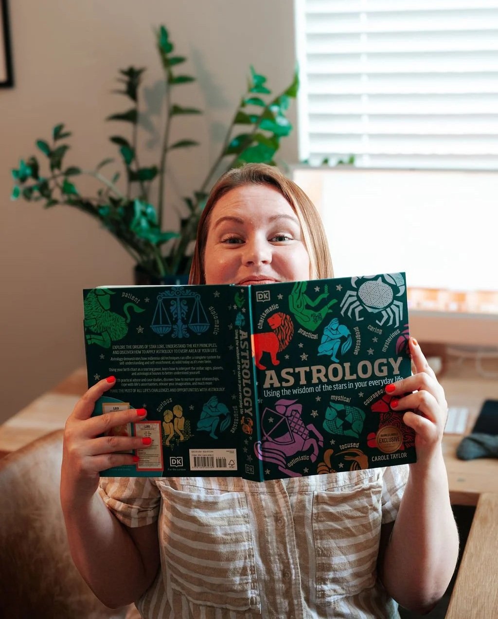 Brunette woman holding an astrology book titled "Astrology: Using the wisdom of the stars in your everyday life" over part of her face with a plant in the background.