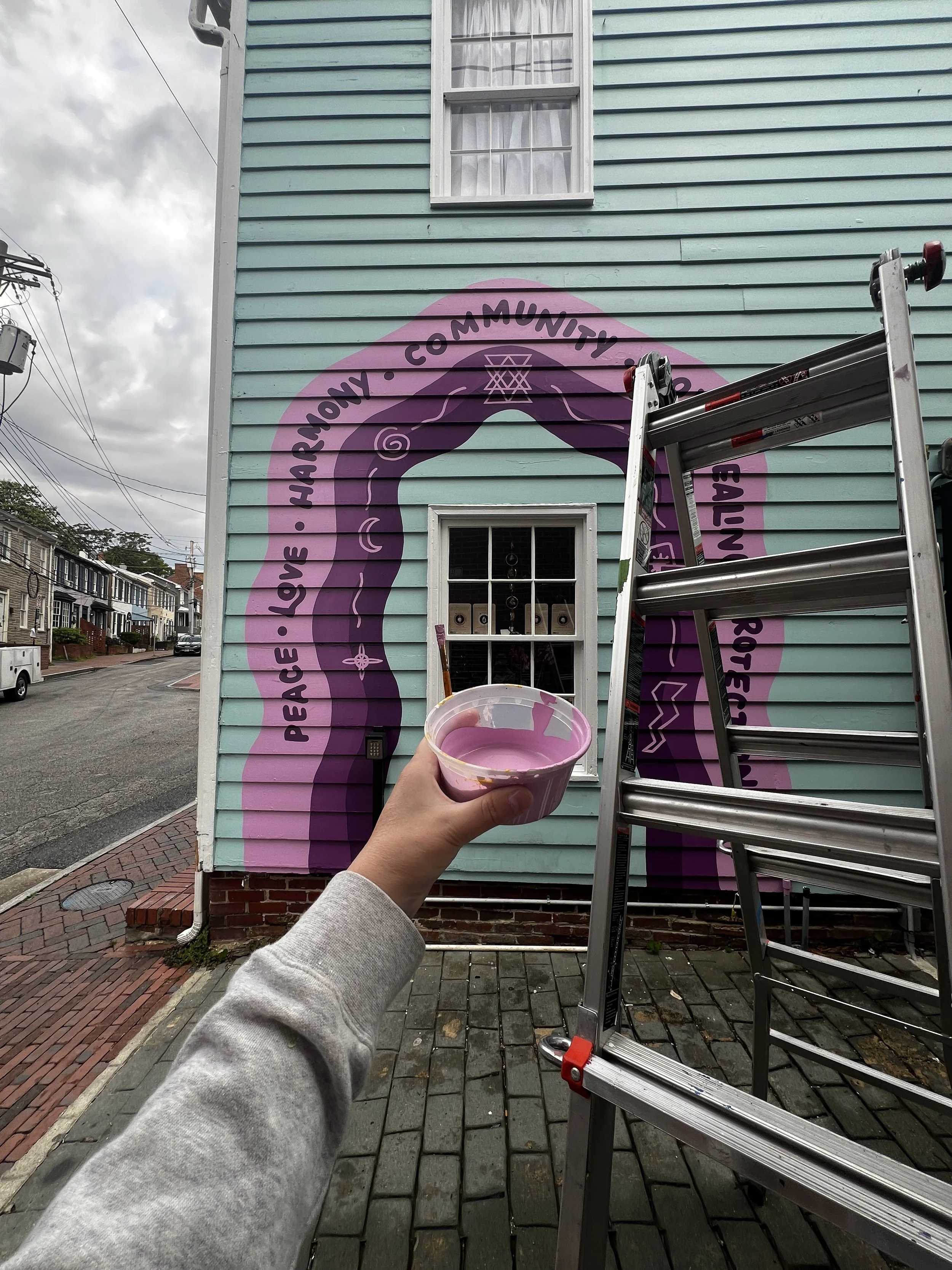 Person holding a cup of pink paint in front of a painted house with a purple circle and black text that reads 'PEACE LOVE CHARITY COMMUNITY EQUALITY BEING REAL'. A ladder leans against the house, which has teal siding and a white window, with other h