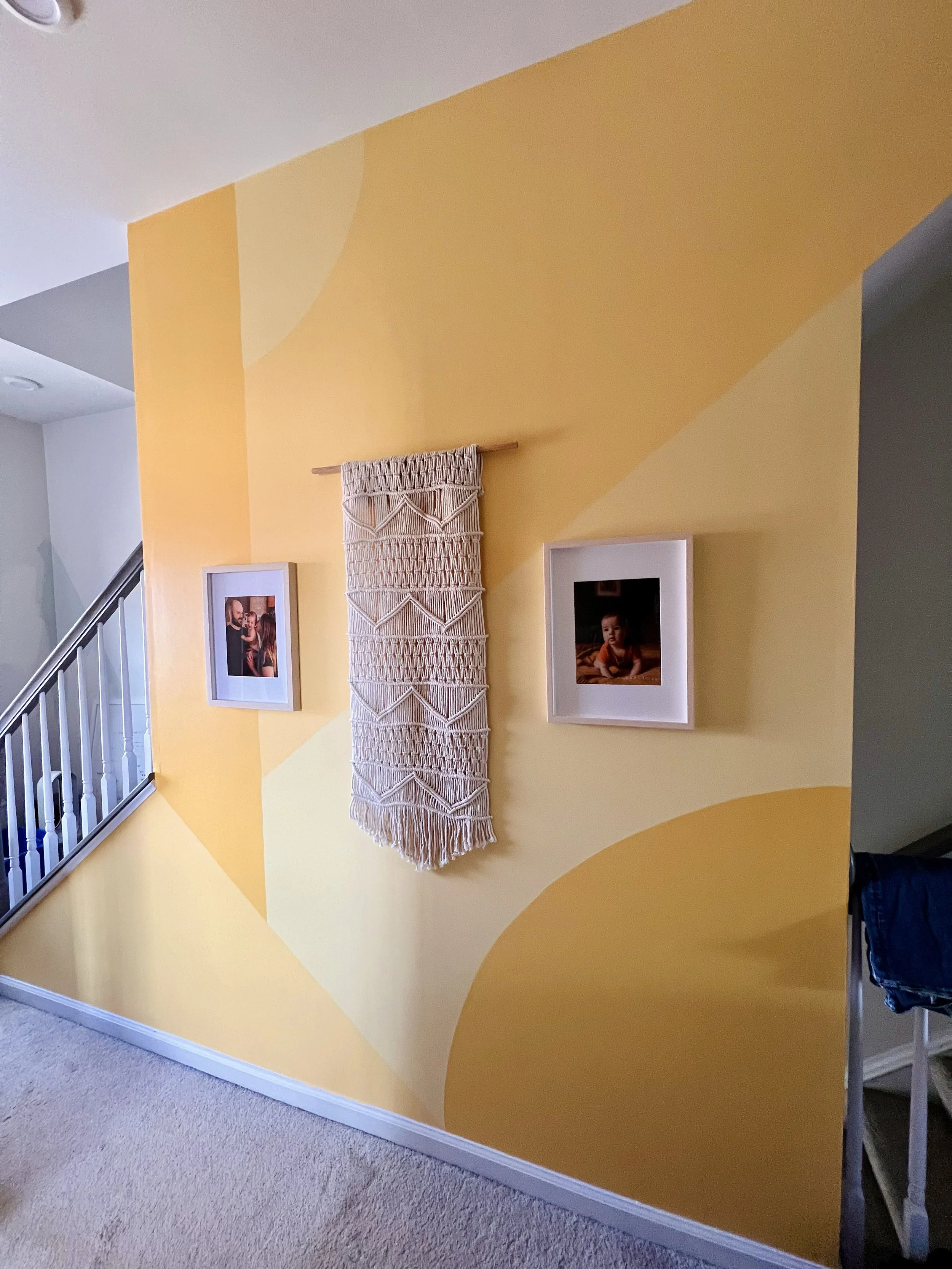 Interior of a home with a yellow wall featuring geometric painted shapes, framed photographs, and a decorative macramé wall hanging above the stair railing.