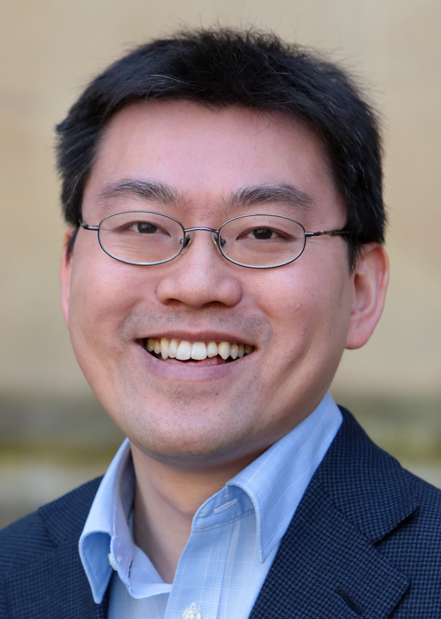 Head & shoulders image of Mark smiling at the camera. He is wearing a dark blue suit jacket and a light blue shirt. He has short dark hair and is wearing glasses with thin metal frames.