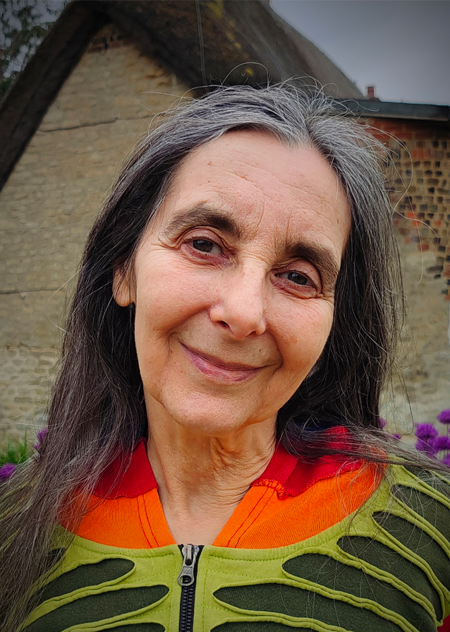 Head & shoulders image of Agnes smiling at the camera. She has long dark and grey hair and is wearing a red, orange, light and dark green zip up top.