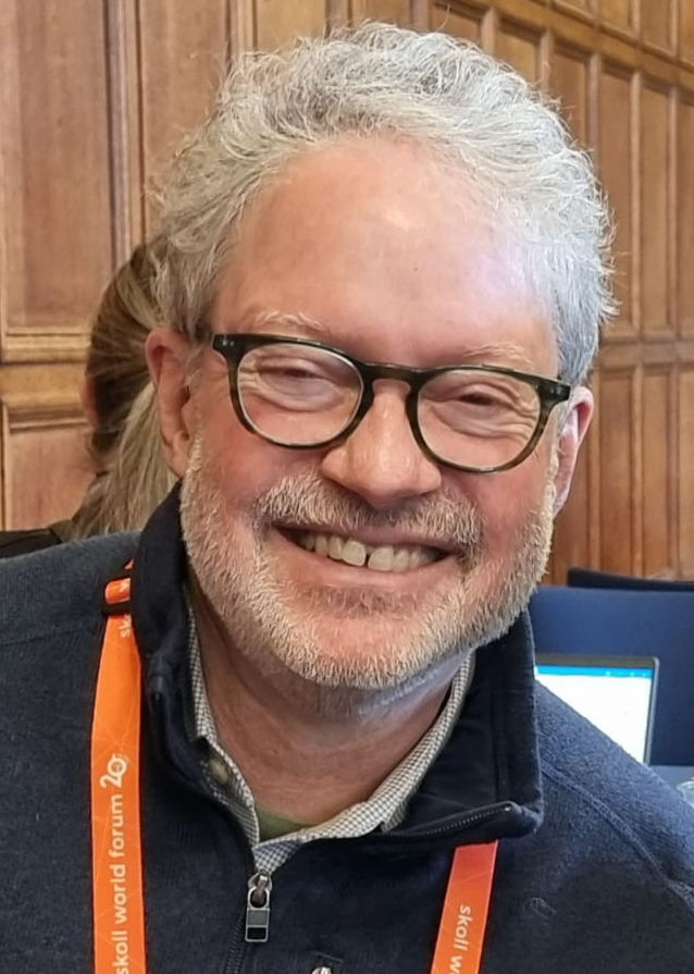 Head and shoulders portrait image of Marc smiling straight at the camera. He is wearing a dark blue jumper, a checked shirt and glasses which have dark brown frames. He has grey hair and a short grey beard.