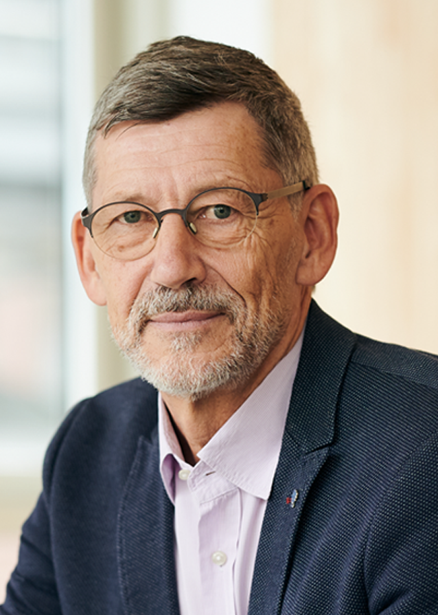 Head and shoulders portrait image of Attila looking straight at the camera. He is wearing a dark blue suit jacket, a light pink, off-white shirt and glasses which have dark grey frames. He has short black and grey hair and a short black & grey beard