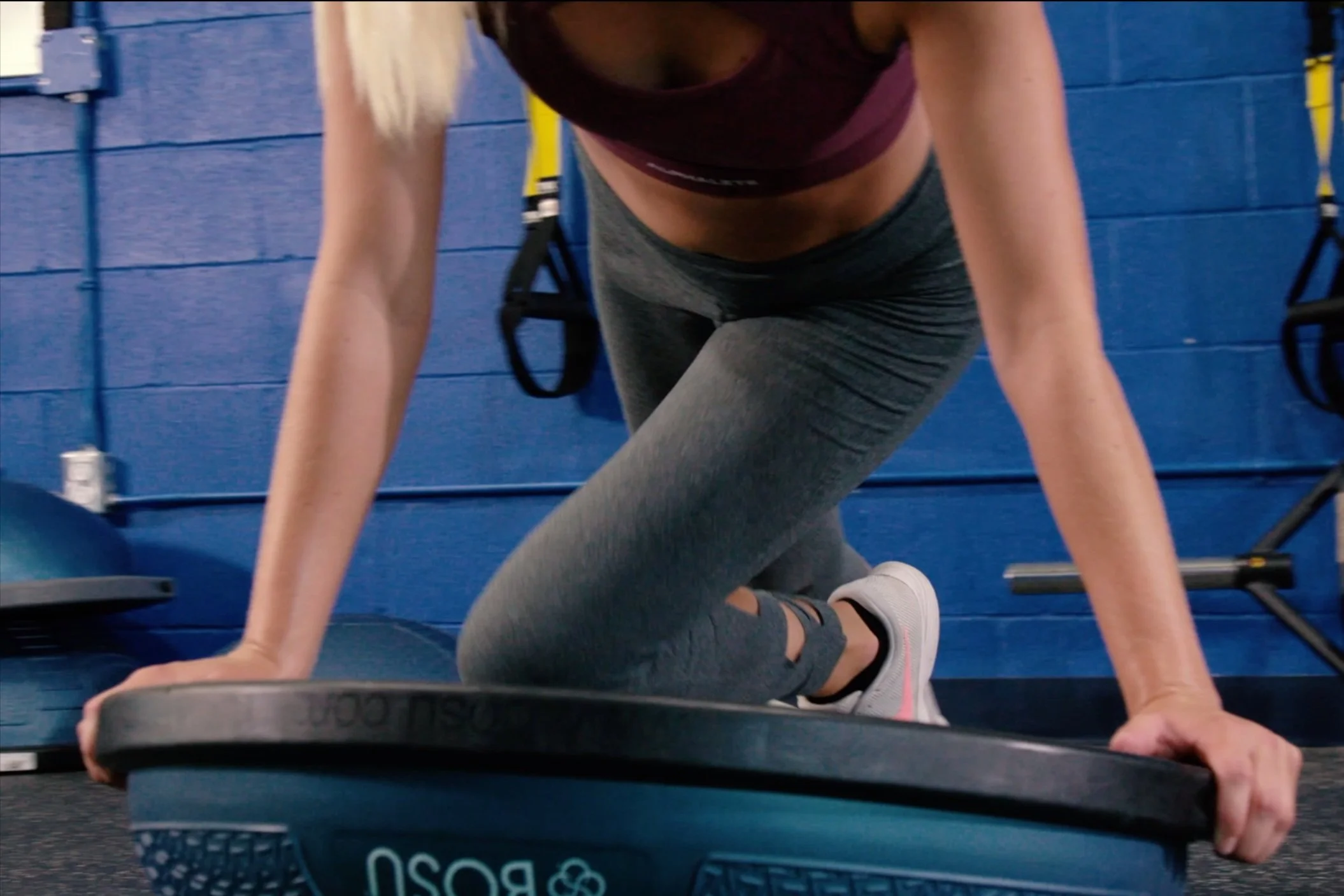 A woman in workout attire, kneeling on a balance board in a gym, with blue walls and fitness equipment in the background.