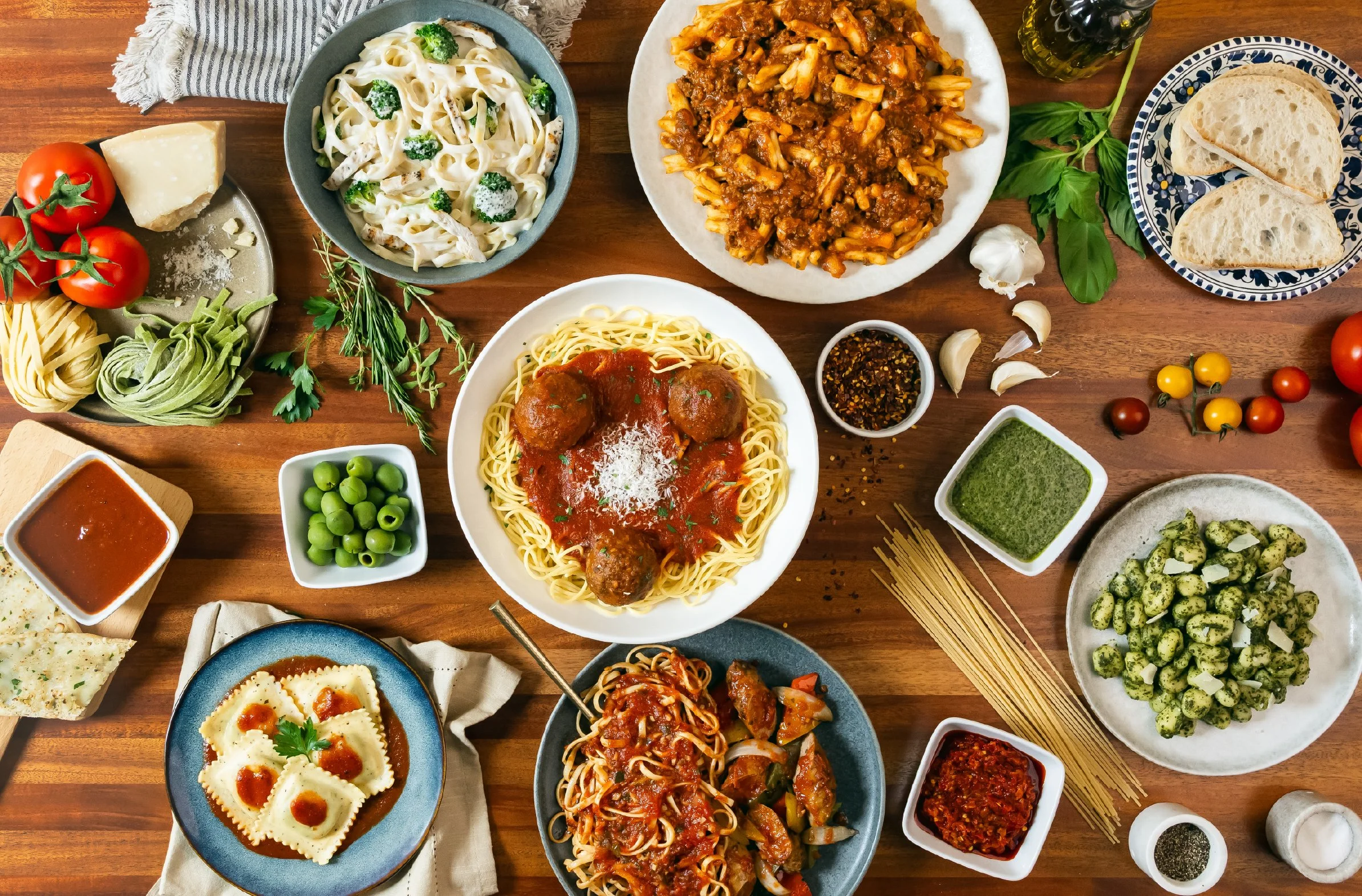 A variety of Italian pasta dishes, including spaghetti with meatballs, feta cheese ravioli, pasta with tomato sauce, and creamy pasta with broccoli, along with assorted vegetables, herbs, and sauces on a wooden table.