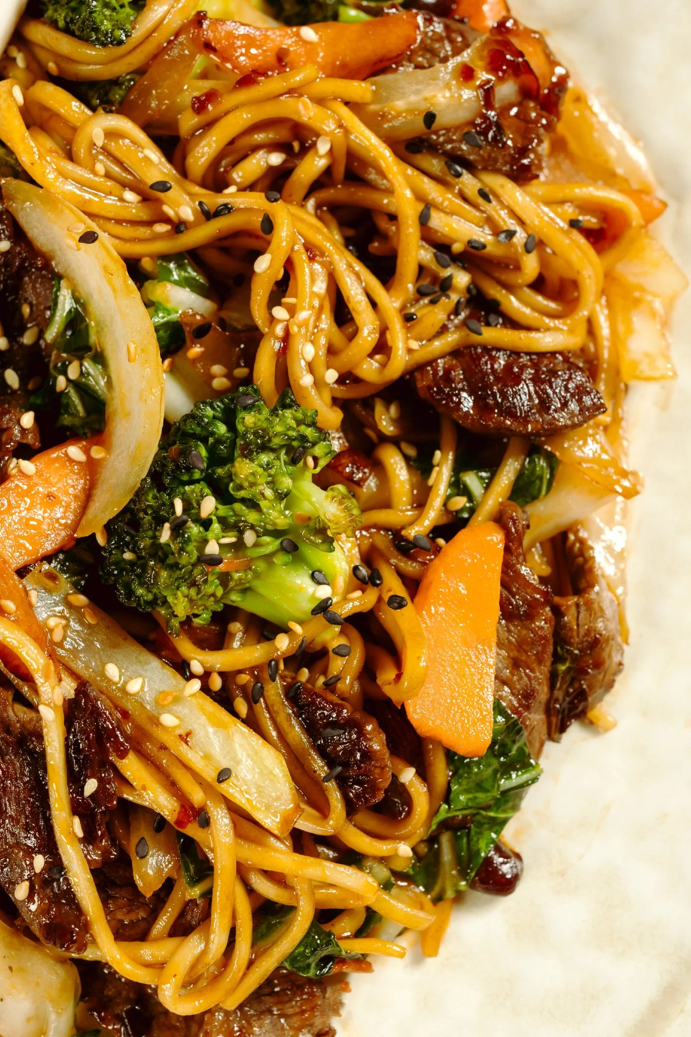 Close-up view of a plate of stir-fried noodles with vegetables, sesame seeds, and pieces of beef.