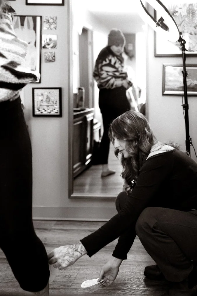 A woman crouching down, adjusting someone's shoe, with a mirror and framed pictures on the wall behind her in a well-lit room.