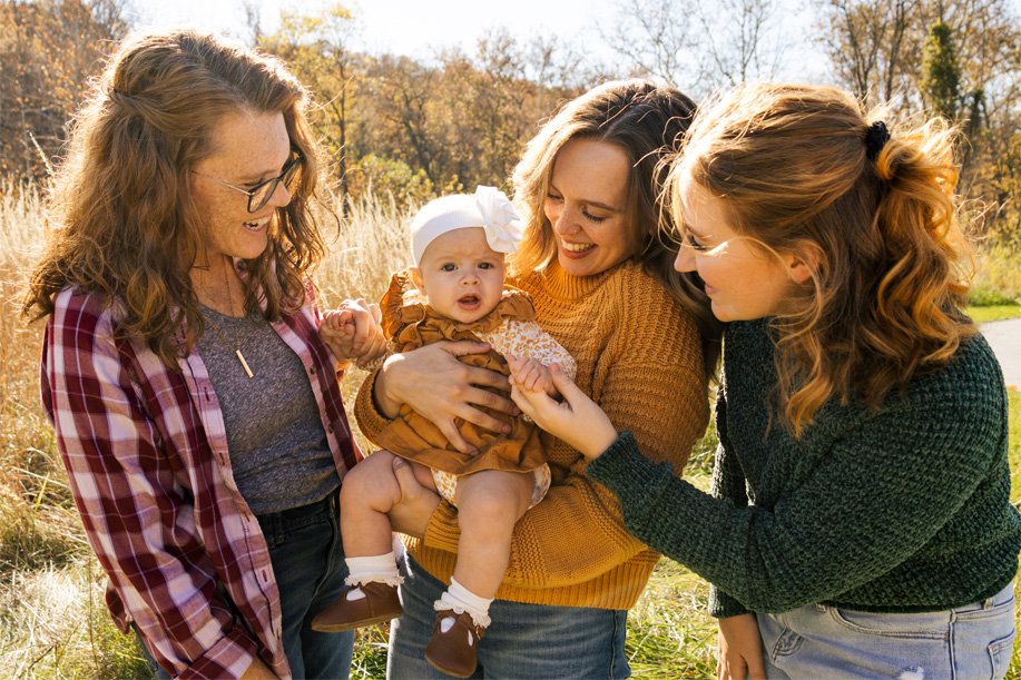 Three women smiling and holding a baby girl outdoors in a sunny field with trees in the background.