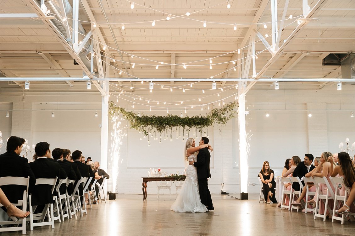 Couple dancing at a wedding reception in a decorated industrial-style venue with string lights and greenery overhead, guests seated on both sides watching.