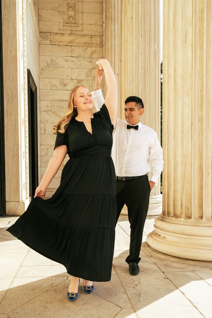 A couple dancing outdoors in front of large classical columns; the woman is wearing a black dress and blue heels, and the man is dressed in a white shirt with a bow tie and black pants.
