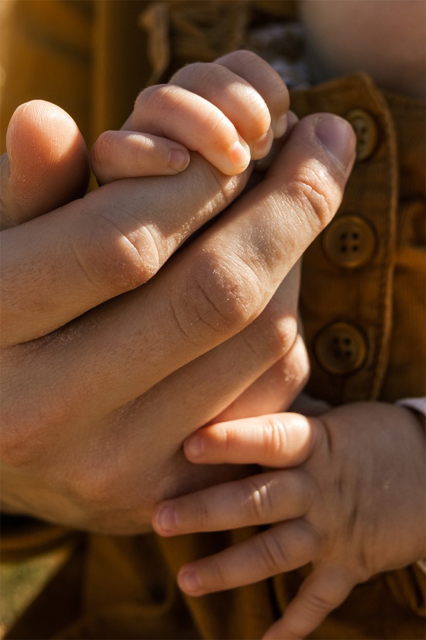 Close-up of an adult's hand holding a child's hand, with a background of a brown jacket with buttons.