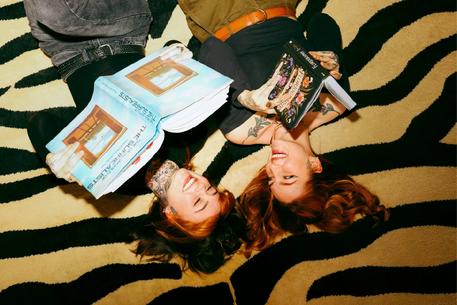 Two women with red hair lying on a black and gold striped carpet, smiling and holding books, with one woman wearing tattoos on her arms.