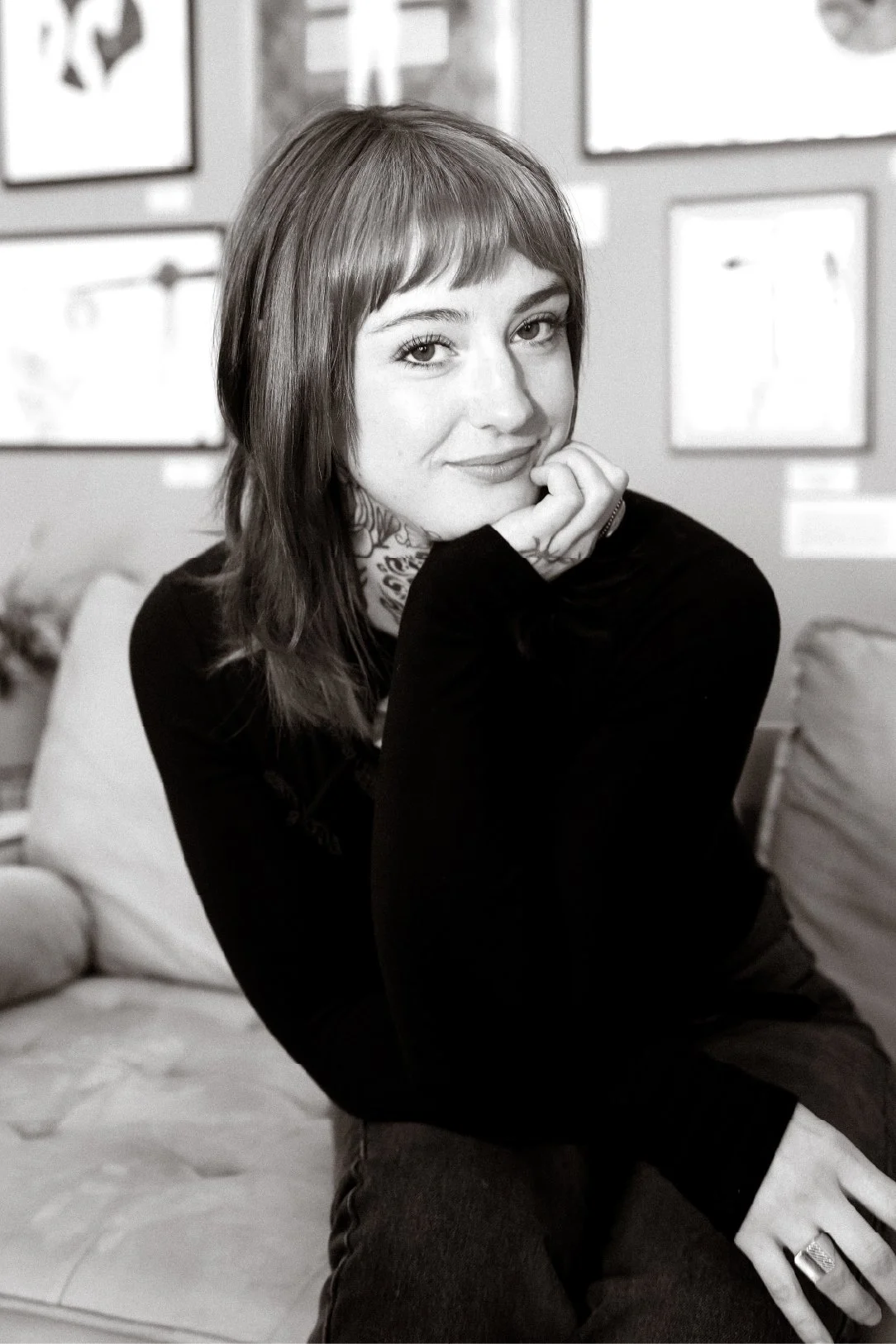 A black-and-white photo of a young woman with shoulder-length hair and bangs, sitting on a couch with artwork on the wall behind her, smiling and resting chin on hand.