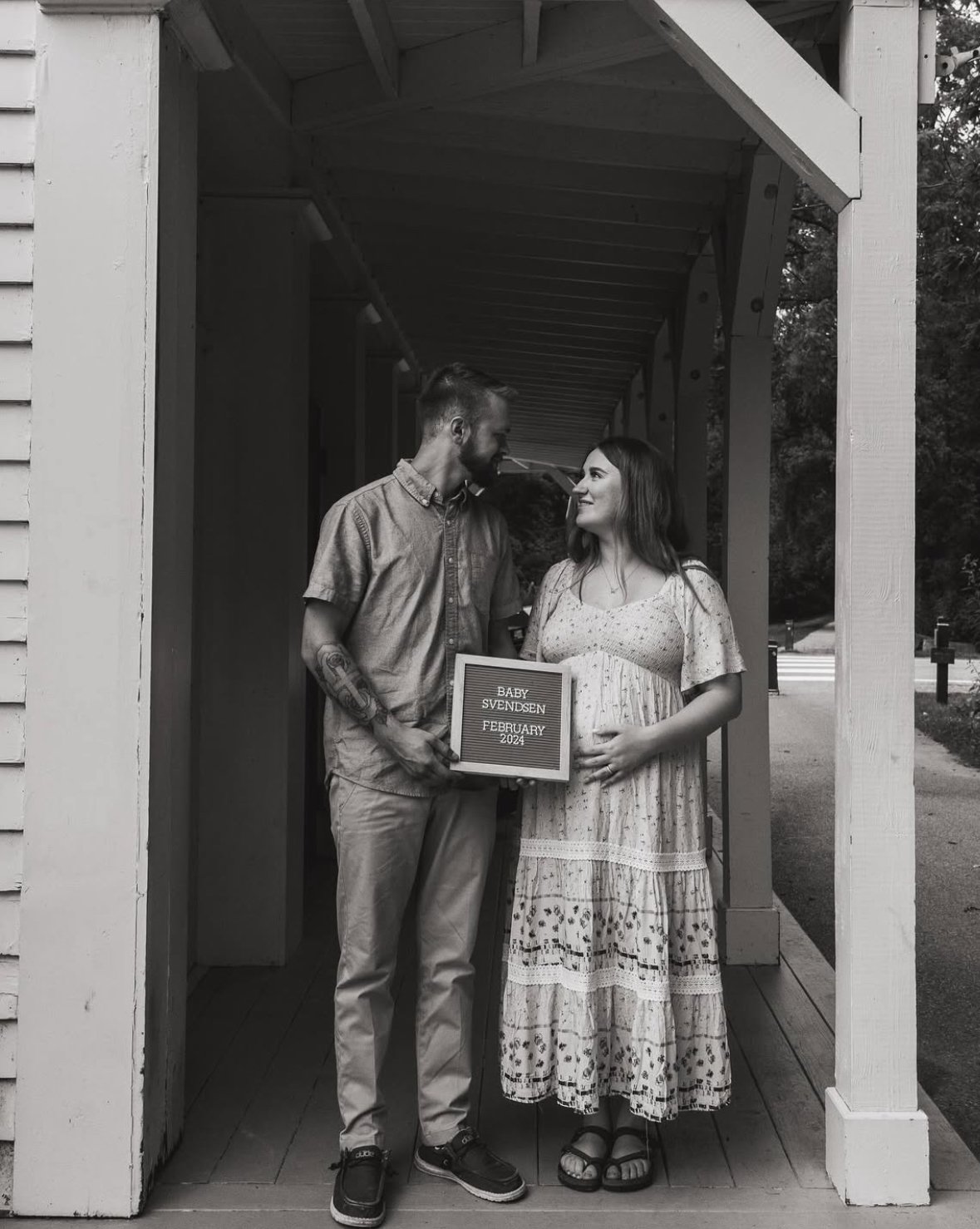 A couple standing in a small outdoor shelter, holding a sign that reads "Baby Svendsen February 2024," with the man leaning close to the woman in a loving moment.