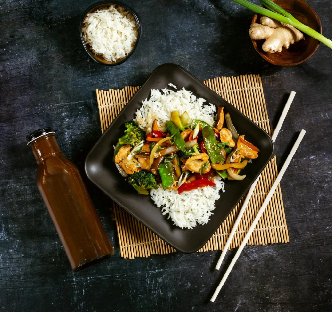 Vegetable stir-fry with chicken served on a black square plate, accompanied by white rice, chopsticks, and a small bowl of rice on a dark surface with a wooden mat.