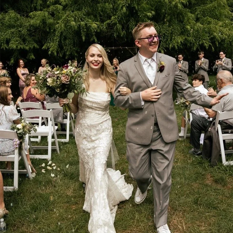 A newly married couple walking arm-in-arm at an outdoor wedding reception, surrounded by friends and family, with lush green trees in the background, the bride holding a large bouquet of flowers and both smiling happy.