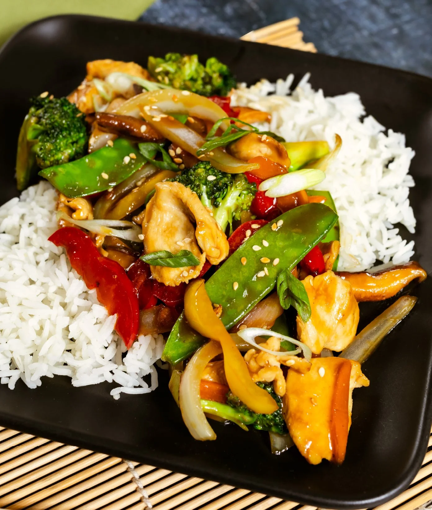 A black bowl filled with white rice and stir-fried vegetables including broccoli, snow peas, red bell peppers, onions, and carrots, garnished with sesame seeds.