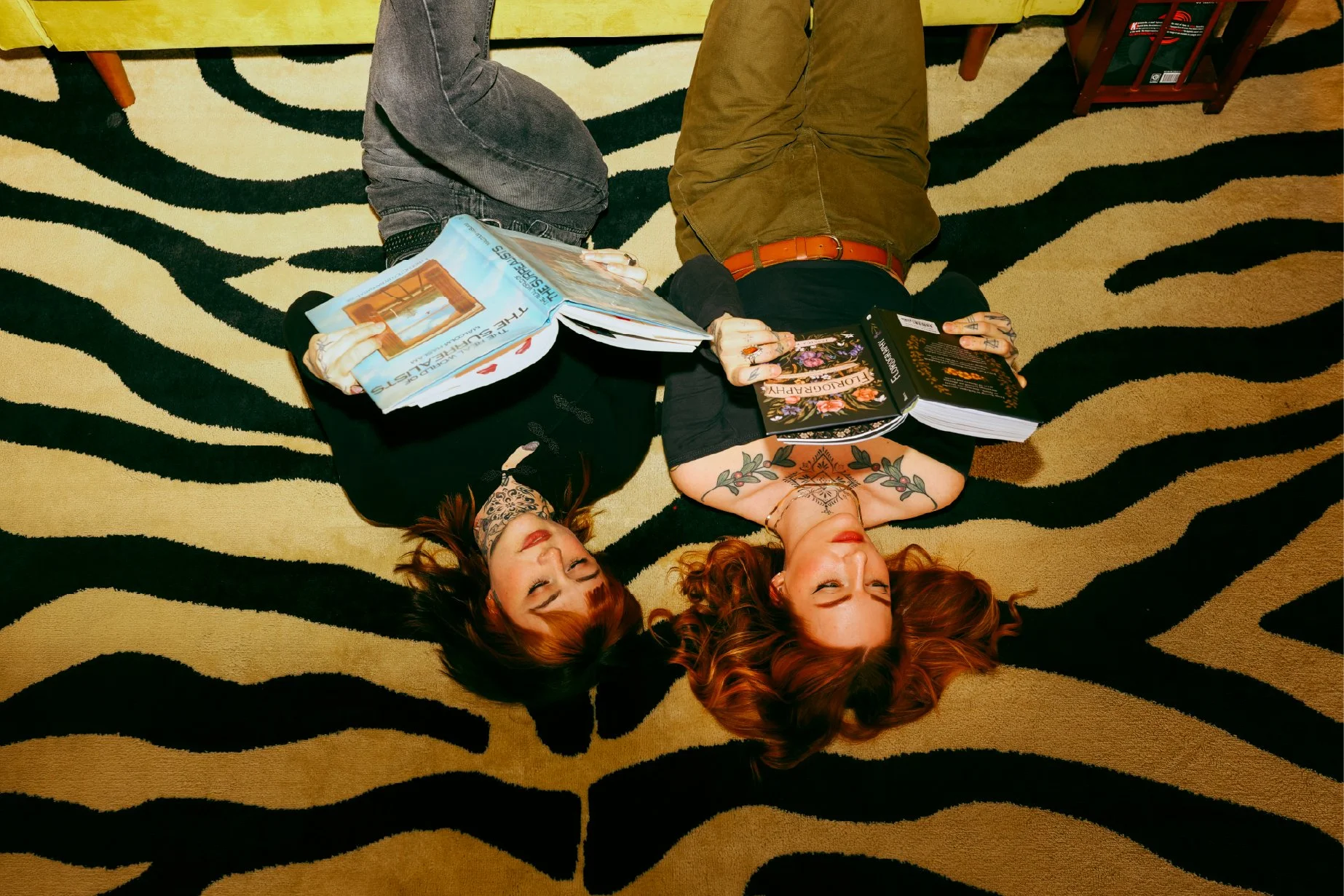 Two women with tattoos lying on a black and beige zebra-patterned carpet, facing up, with books resting on their bodies.