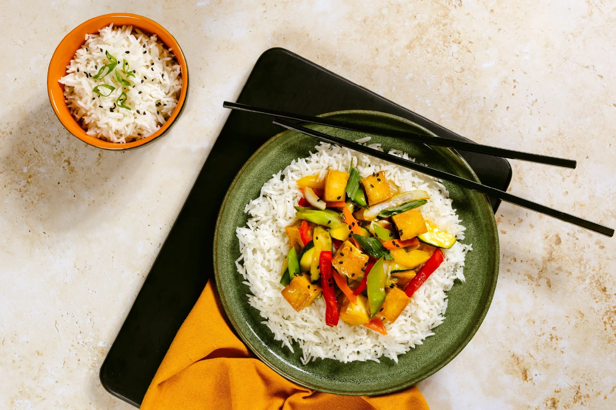 A plate of stir-fried vegetables and tofu on a bed of white rice, with a bowl of rice on the side, chopsticks resting on the plate, on a black tray with a yellow and orange napkin beneath.