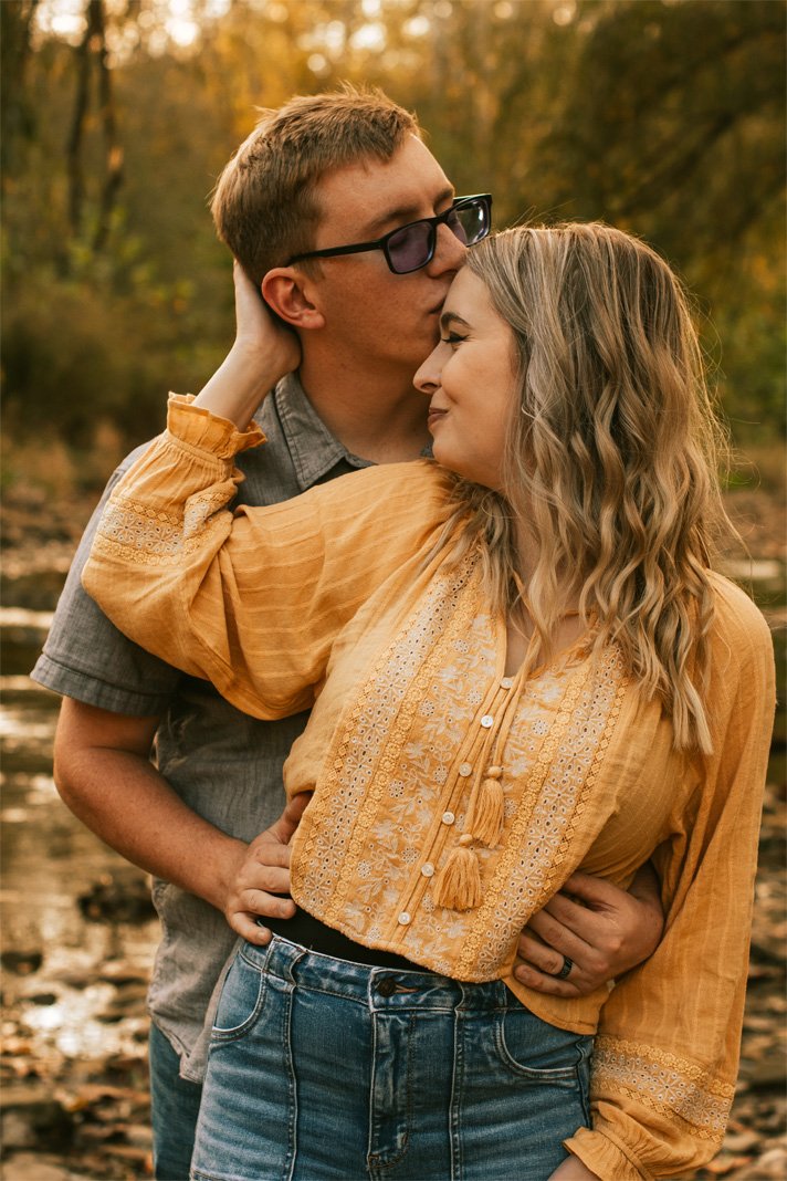 A couple embracing outdoors, with the man kissing the woman's forehead, surrounded by fall trees and warm lighting.