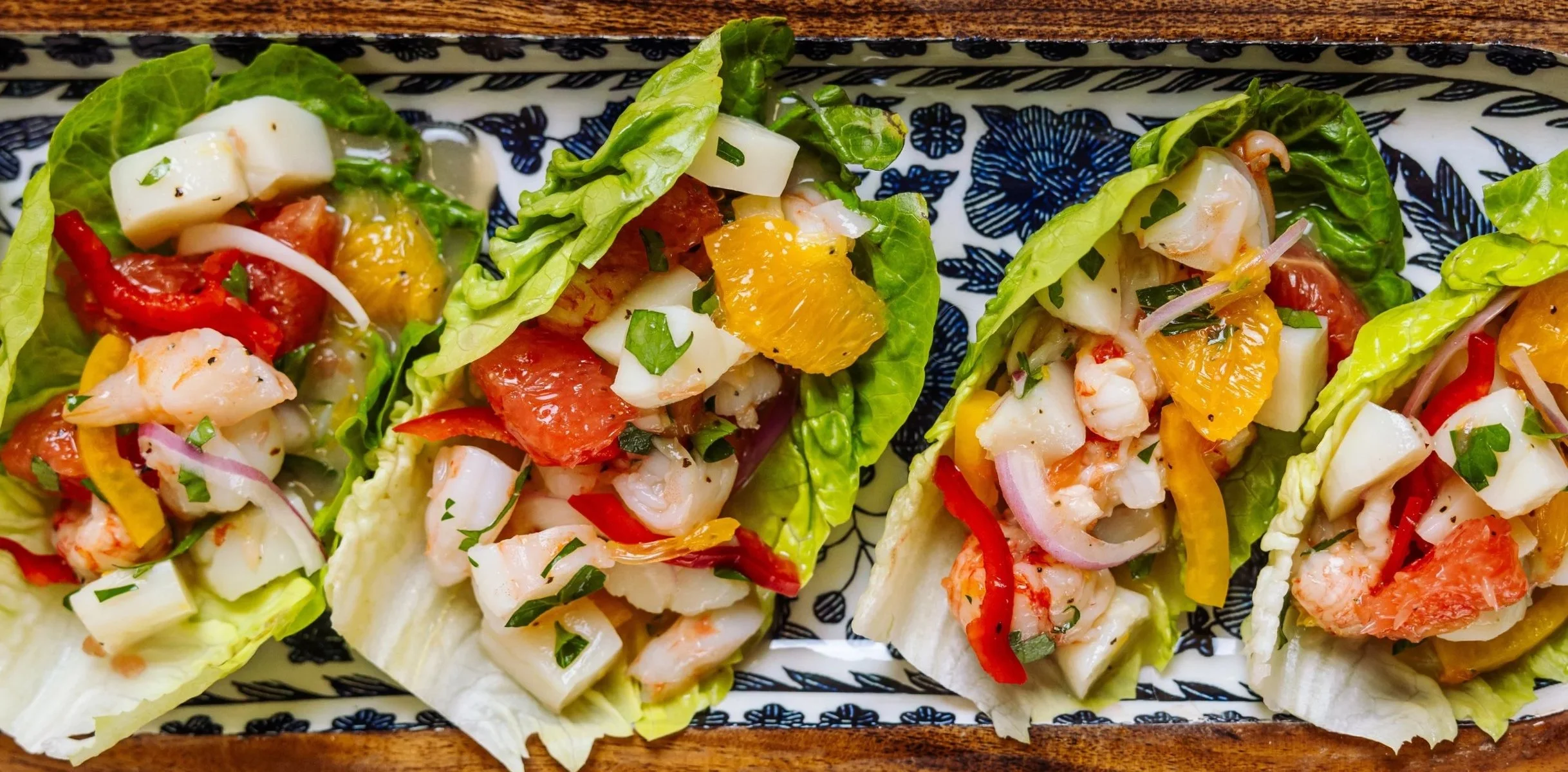 Close-up of four lettuce cups filled with chopped seafood, diced vegetables, and herbs, on a decorative rectangular tray with black and white floral patterns, on a wooden table.