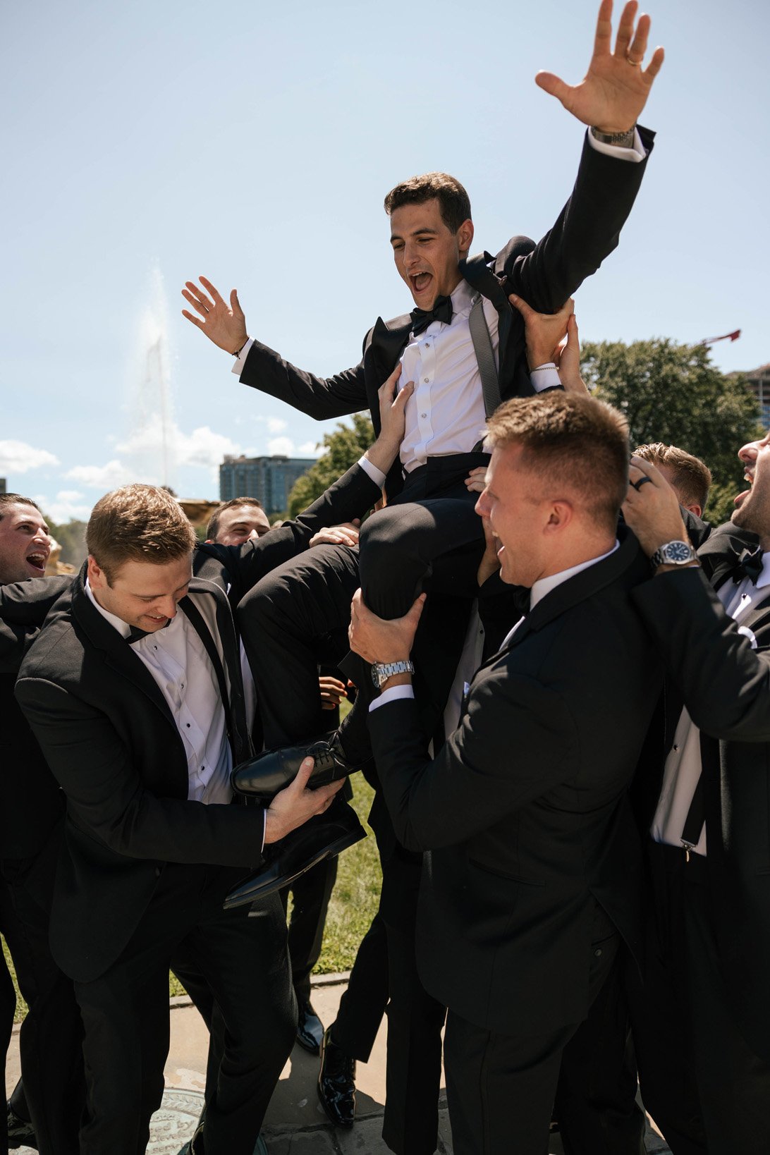 A group of men dressed in tuxedos lifting a man in a tuxedo on their shoulders outdoors on a sunny day, celebrating and smiling.