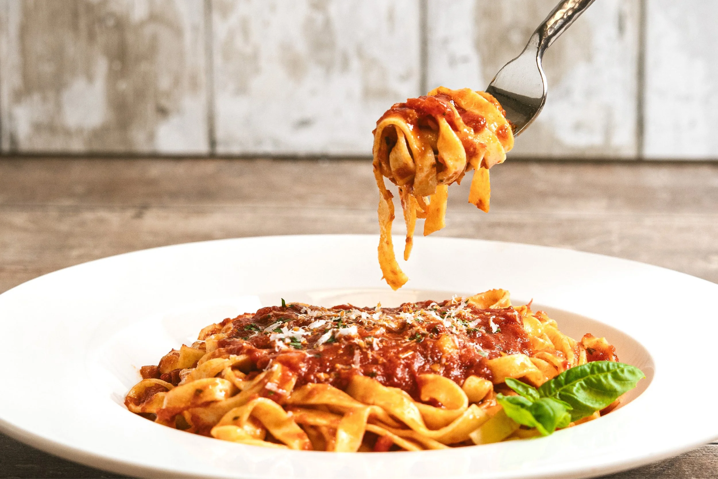 A fork lifting a portion of spaghetti with meat sauce above a white plate containing more spaghetti and a basil leaf, set against a rustic background.
