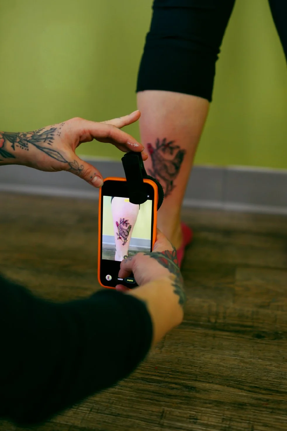 Person with tattoos is using a smartphone to take a picture of their calf tattoo, which is visible in the foreground. The person is standing on a wooden floor with a green wall in the background.