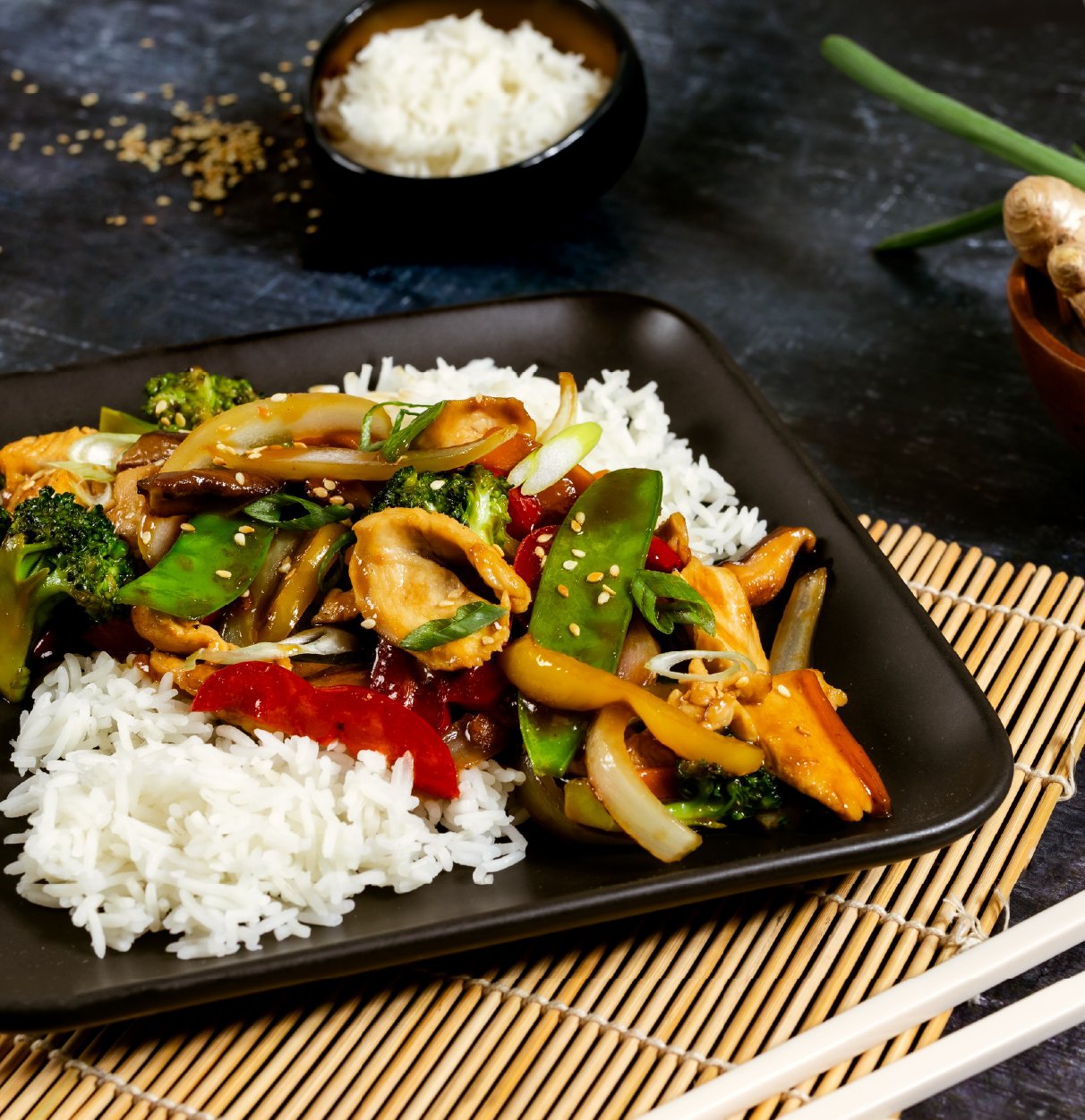 Stir-fried vegetables with chicken served over white rice on a black plate.