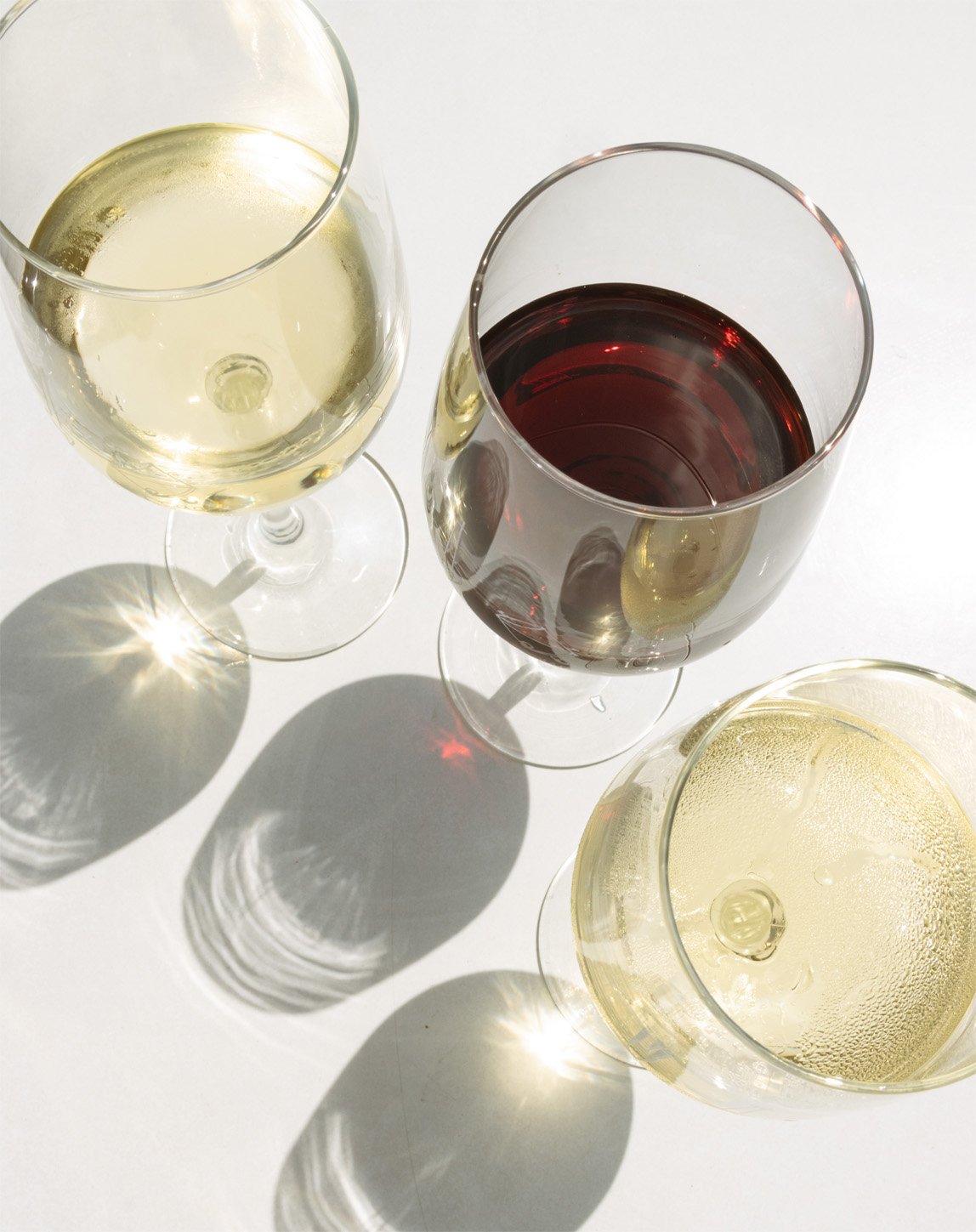 Three wine glasses on a white surface, two with white wine and one with red wine, casting shadows and reflections.