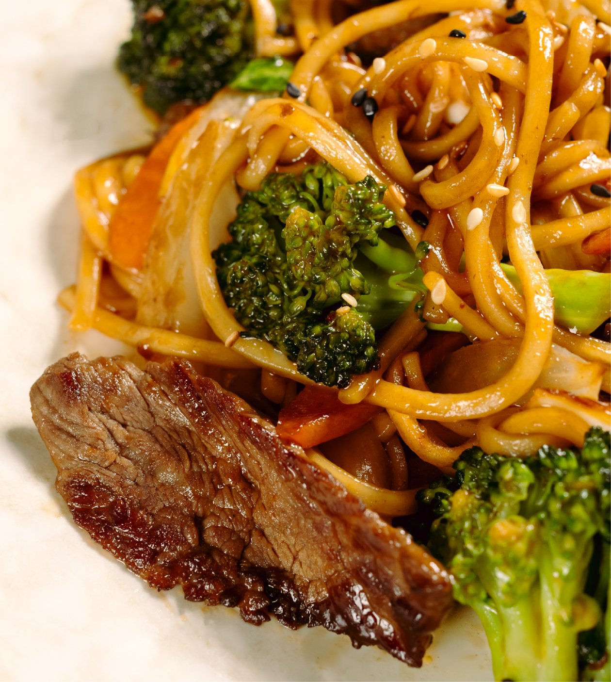 Close-up of Asian-style stir-fried noodles with broccoli, carrots, and sesame seeds, with a piece of cooked beef.