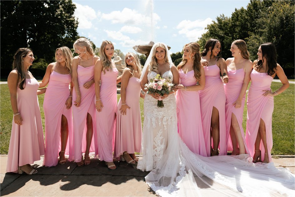 Bride in a white lace wedding dress with veil holding a bouquet among her bridesmaids in pink dresses outdoors on a sunny day.