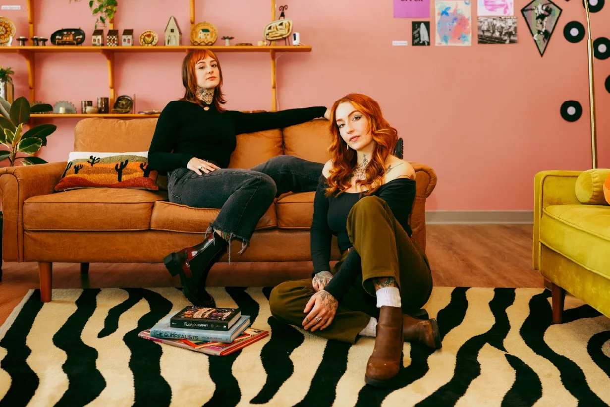 Two women with red hair in a colorful, cozy living room. One is sitting on a sofa with her arm resting on the back, and the other is sitting on the floor with her legs crossed. The room has pink walls, decorative shelves with various objects, and a z